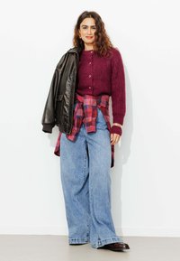 Pull en maille côtelée bordeaux avec des boutons, jean large en denim, chemise à carreaux nouée à la taille, et une veste en cuir noire posée sur l'épaule.