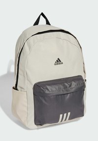 Zaino beige con tasca frontale grigia, zip nera e logo Adidas. Presenta una maniglia superiore e spallacci imbottiti per il comfort.