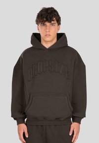 Giovane uomo che indossa una felpa oversize marrone scuro con tasca frontale e testo in rilievo "Dropsize" sul petto, contro uno sfondo semplice.