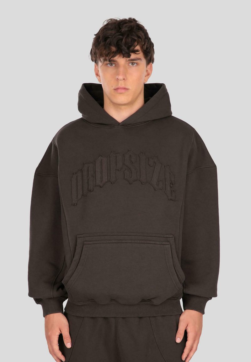 Giovane uomo che indossa una felpa oversize marrone scuro con tasca frontale e testo in rilievo "Dropsize" sul petto, contro uno sfondo semplice.