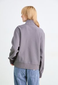 Persona con cabello rubio que viste un sudadera de cuello alto gris y vaqueros azules holgados, de pie de espaldas frente a un fondo simple.