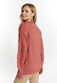Maglione color ruggine con maniche testurizzate e polsini a coste. Presenta dettagli floreali sulle maniche, con una vestibilità ampia e colletto a girocollo.