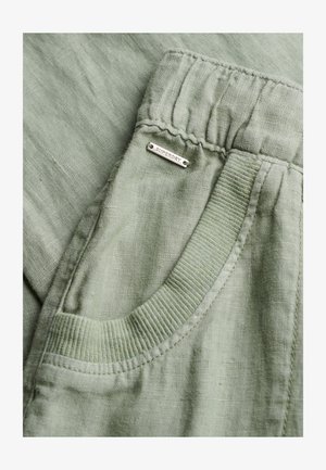 Pantalones de lino verde claro con cinturilla elástica y bolsillo con borde acanalado, con una pequeña placa metálica etiquetada como "SUPERDRY" cerca del bolsillo.