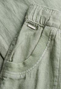 Pantalon en lin vert clair avec taille élastique et poche à bord côtelé, comportant une petite étiquette métallique portant l'inscription "SUPERDRY" près de la poche.