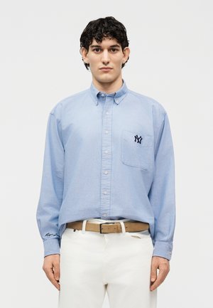 Άνδρας που φοράει ανοιχτό μπλε πουκάμισο με σκούρο λογότυπο στην τσέπη του στήθους, μπεζ ζώνη και λευκό παντελόνι, στέκεται μπροστά σε λευκό φόντο.