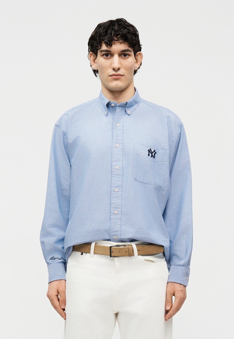 Άνδρας που φοράει ανοιχτό μπλε πουκάμισο με σκούρο λογότυπο στην τσέπη του στήθους, μπεζ ζώνη και λευκό παντελόνι, στέκεται μπροστά σε λευκό φόντο.