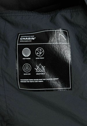 Zwarte stoffen label met het "CHASIN'" logo, onderhoudsinstructies en iconen voor thermisch en vochtafvoerend beheer. Tekst is vetgedrukt met witte accenten.