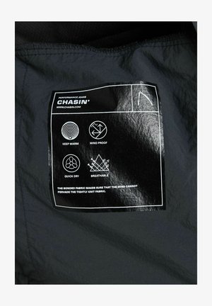 Musta kangasriippu, jossa on "CHASIN'" logo, hoito-ohjeet ja ikonit lämpö- ja kosteusjohtamiseen. Teksti on lihavoitua ja valkoisilla korostuksilla.