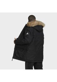 Černá parka s kapucí vyplněnou kožešinou, polstrovaný design, přední kapsy a logo Adidas na levém rukávu. Nastavitelné manžety.