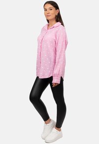 Camicia rosa a maniche lunghe con motivi a cuore bianchi, vestibilità rilassata, chiusura a bottoni sul davanti, abbinata a leggings neri e sneakers bianche.