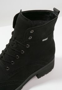 Bottine en daim noir avec lacets à l’avant, détails brogue décoratifs, œillets métalliques et petit talon empilé.