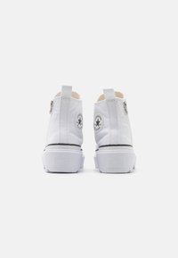 Białe płótna buty typu high-top z zamkiem błyskawicznym, grube gumowe podeszwy oraz okrągłe naszywki z logo na zewnętrznej części pięty i języku.
