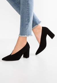 Persona che indossa jeans azzurri con orlo sfrangiato e décolleté neri in suede con tacco a blocco, in piedi su un pavimento bianco.
