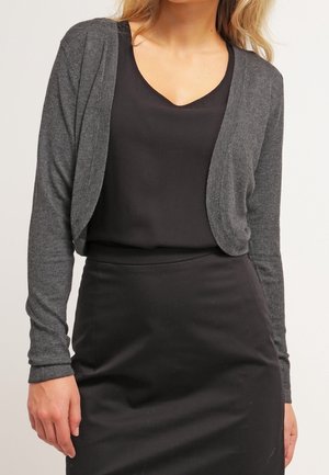 Femme portant un haut noir à col en V, une jupe noire et un cardigan court gris foncé ouvert sur le devant.