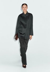 Donkergrijze mantel met hoge kraag en twee zijzakken, gecombineerd met bijpassende donkere broek en een bruine tas, met een soepele stofstructuur.