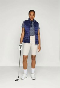 Námornícky prešívaný vest v topánke so vzorovaným modrým polo tričkom, krémovými šortkami a bielymi topánkami. Drží golfovú palicu, stojí na neutrálnom pozadí.