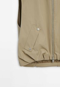 Veste beige sans manches avec fermeture éclair à l'avant et poche latérale avec rabat sécurisé par deux boutons-pression métalliques.