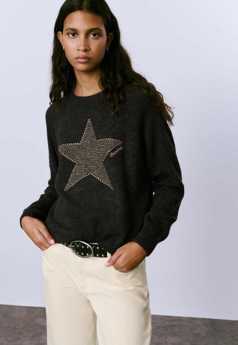 Suéter de punto gris oscuro con una gran estrella beige en el centro, adornada con detalles de tachuelas, combinado con pantalones beige claros y un cinturón negro.