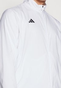 Veste blanche à fermeture éclair fabriquée en matériau léger, avec un col montant, un logo noir sur la poitrine et des accents perforés sur les manches.