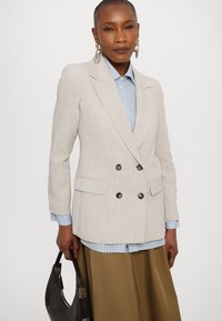 Lys beige dobbelbrystet blazer med strukturerte skuldre, svarte knapper og to frontlommer. Bæres over en lyseblå rutet skjorte.