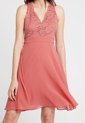 Vrouw draagt een mouwloze roze jurk met een kanten lijfje en een vloeiende rok, staand tegen een effen lichte achtergrond.