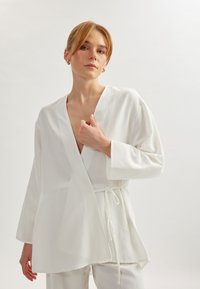 Top blanco cruzado hecho de una tela ligera, con un escote en V profundo, mangas largas y cintura con lazo. Textura suave con diseño minimalista.