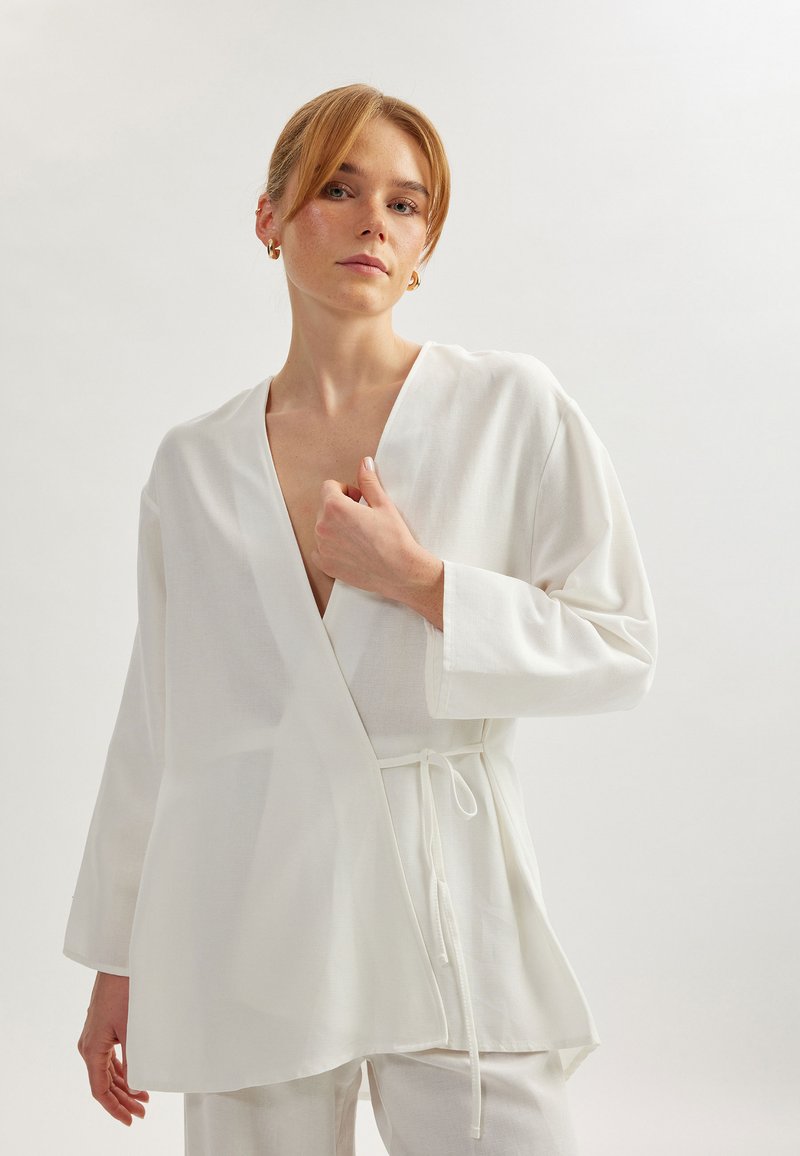 Top blanco cruzado hecho de una tela ligera, con un escote en V profundo, mangas largas y cintura con lazo. Textura suave con diseño minimalista.
