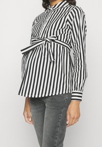 Svart och vit randig gravidblus med knytning i midjan, långa ärmar och ståkrage, kombinerad med grå denimjeans.