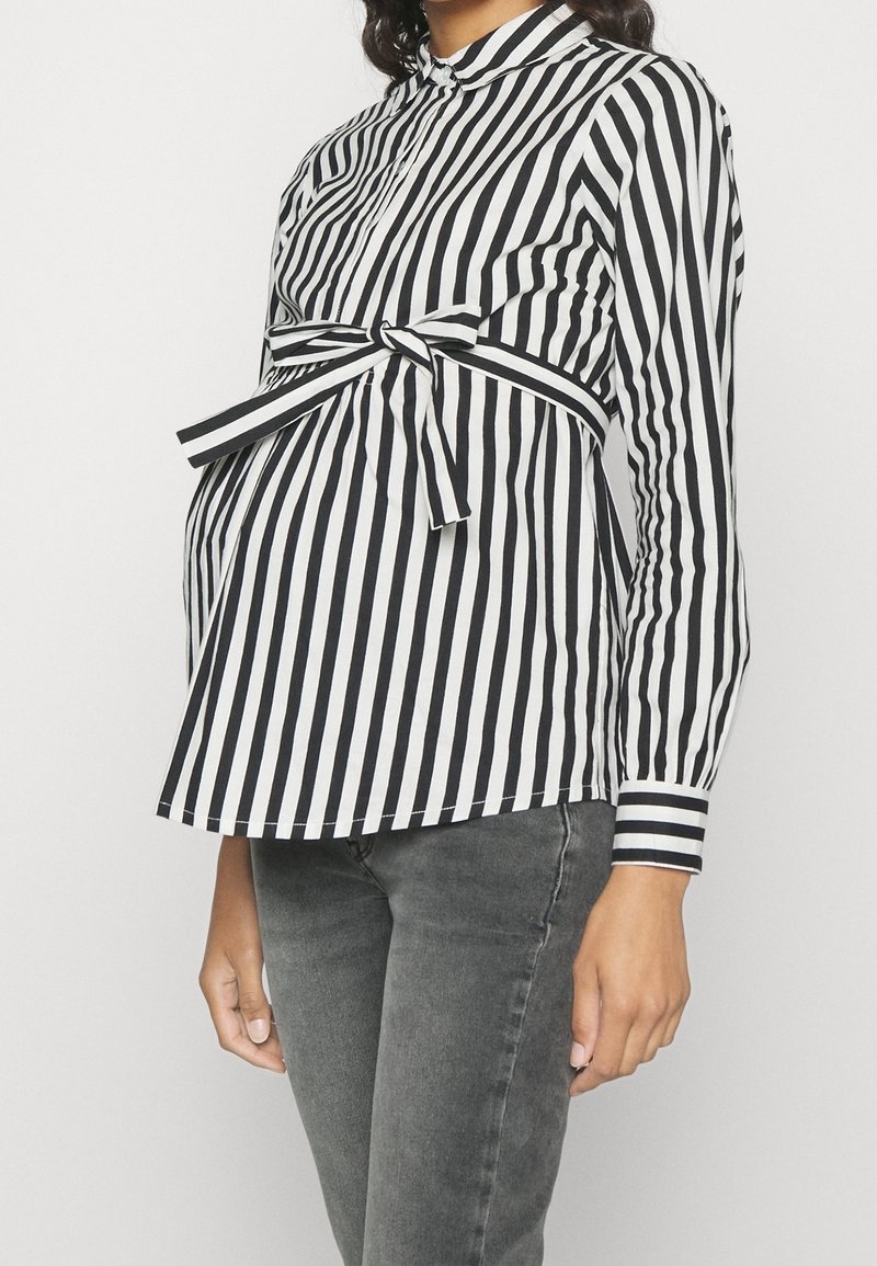 Svart och vit randig gravidblus med knytning i midjan, långa ärmar och ståkrage, kombinerad med grå denimjeans.