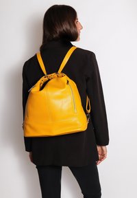 Zaino in pelle gialla con coulisse, dotato di due tasche con zip, forma arrotondata e spallacci regolabili, posizionato su un cappotto nero.