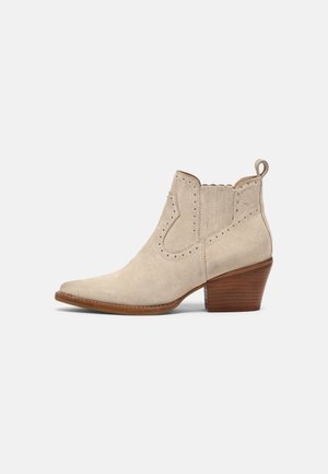 Bottine beige en daim avec petit talon en bois superposé, languette, panneau latéral élastique et détails décoratifs perforés.