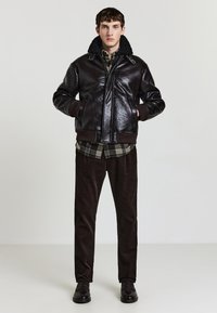 Braune Lederjacke mit Schafswollkragen, kombiniert mit einem karierten Hemd, braunen Cordhosen und schwarzen Slippern. Einfaches Design und Textur.