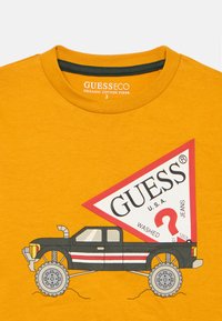 Maglietta gialla con una grafica di un pickup nero e un grande etichetta a forma di triangolo rosso con la scritta "GUESS U.S.A." e "WASHED JEANS."