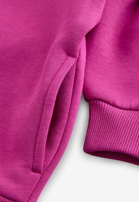 Felpa fucsia con polsini e orlo a coste. Presenta una texture liscia e cuciture in contrasto lungo la cucitura della manica.