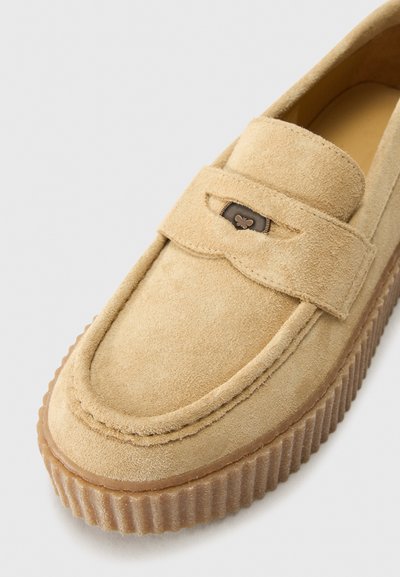 Mocassin en daim beige avec un bout rond, semelle en caoutchouc et une sangle décorative avec un emblème marron. Tige texturée avec des accents cousus.