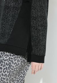 Primer plano de una persona vistiendo una camiseta de manga larga negra con manchas y pantalones gris con estampado de leopardo, de pie contra una pared clara.
