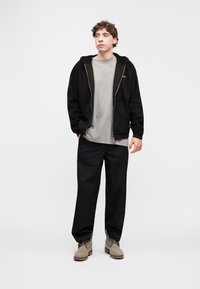 Sweat à capuche en polaire noir avec fermeture éclair devant, t-shirt gris, pantalon large noir, et bottes lacées couleur tan. Tenue simple et décontractée.