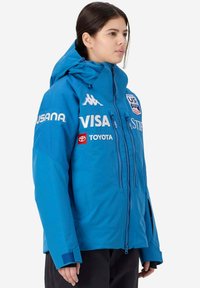 Giacca impermeabile blu con cappuccio regolabile, chiusura con zip e toppe con logo. Presenta polsini elastici e una vestibilità ampia per un abbigliamento a strati.