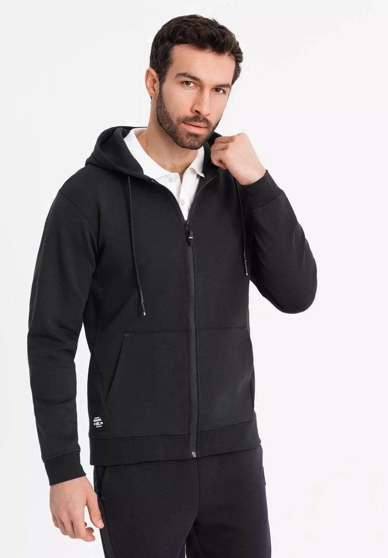 Sort zip-up hoodie med en frontlomme, snørebånd og en glat tekstur. Båret over en hvid skjorte med krave, der har en subtil logo-detalje.