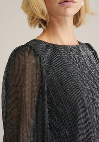 Rochie neagră texturată cu mâneci din material transparent, decorate cu dungi din fir argintiu. Decolteu rotund și pliu subtil pe umeri.