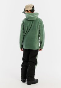 Felpa in pile di colore verde tenue, con cappuccio, maniche lunghe e vestibilità ampia, abbinata a pantaloni neri e stivali robusti.