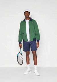 Άνδρας με πράσινο μπουφάν, λευκό καπέλο, σκούρο μπλε σορτς, λευκά αθλητικά παπούτσια, κρατώντας ρακέτα του τένις, στέκεται μπροστά σε απλό λευκό φόντο.