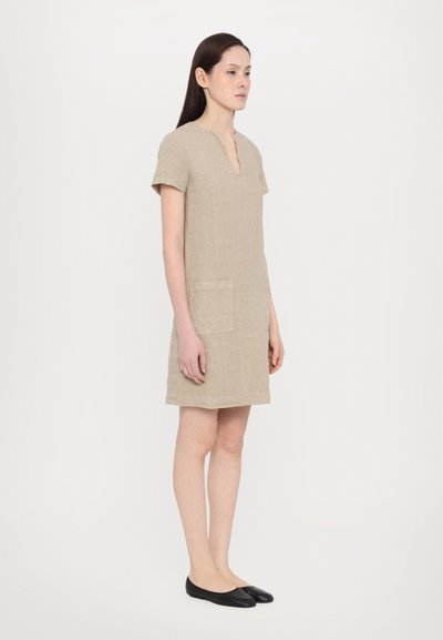 Korte mouwen beige linnen jurk met een V-hals, voorzien van een voorzak en een losse pasvorm, gecombineerd met zwarte ballerina's.