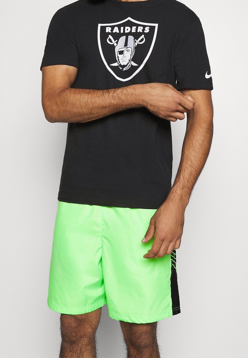 Homme portant un t-shirt noir des Raiders et un short de sport vert fluo, debout devant un fond uni.