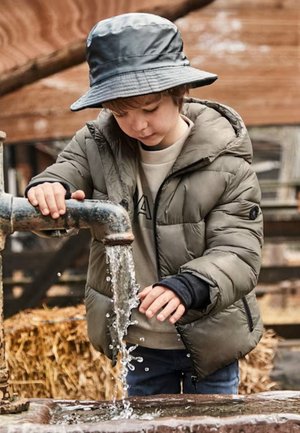Giacca imbottita verde oliva con cappuccio, camicia blu e jeans scuri. Le mani di un bambino girano un rubinetto, l'acqua scorre in una bacinella di legno.
