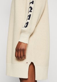 Abito maglione in un beige chiaro con polsini a coste, dotato di spacchi laterali e un dettaglio logo blu e nero sulla manica.