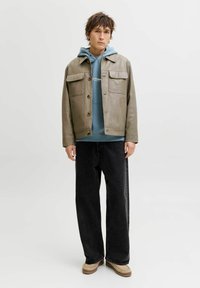 Giacca in pelle grigio chiaro con due tasche sul petto, indossata sopra una felpa blu con cappuccio. Pantaloni neri a gamba larga e scarpe beige slip-on.