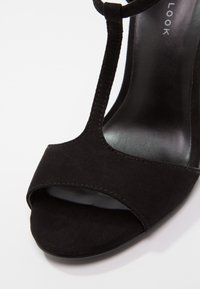 Sandale à talons hauts noirs en daim. Présente un design à lanière T, une ouverture au niveau des orteils et une semelle intérieure noire lisse. La forme courbée du talon ajoute de l'élégance.