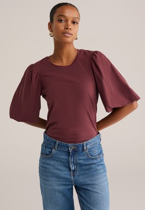 Haut maroon à manches courtes avec des manches bouffantes, associé à un jean en denim bleu taille haute. Texture douce et détail de col rond.