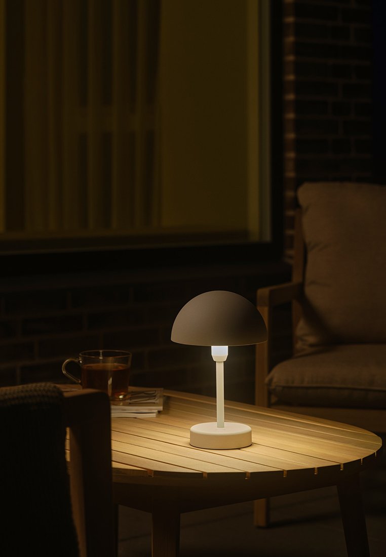 Tischlampe mit einem runden grauen Schirm, weißem Stamm und runder Basis auf einem Holztisch, beleuchtet in einer schwach beleuchteten Außenumgebung.